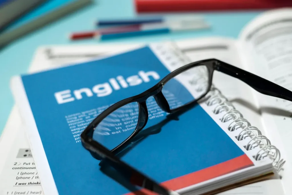 Óculos de leitura pousados sobre um livro azul com a palavra "English" na capa, aberto em uma página com texto e exercícios, simbolizando o estudo da gramática inglesa.