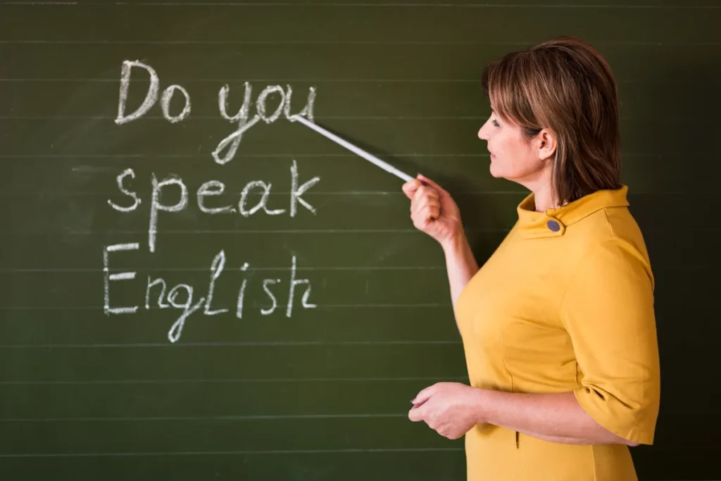 Professora de inglês em frente a um quadro-negro com a frase "Do you speak English?" escrita em giz, apontando para a gramática inglesa.