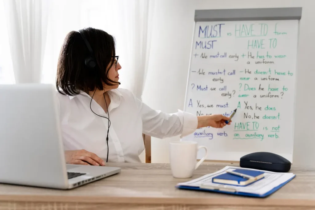 Professora com fone de ouvido em frente a um quadro branco com regras de gramática inglesa ("MUST vs HAVE TO"), ensinando online com laptop e materiais de estudo.