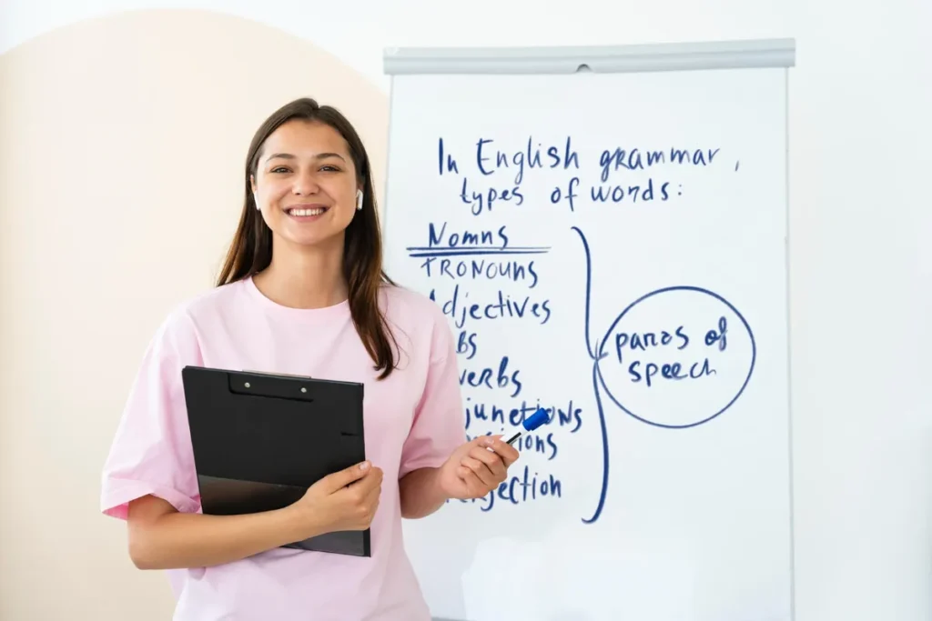 Jovem professora sorridente explicando gramática inglesa e tipos de palavras (parts of speech) em um quadro branco. A professora usa um fone de ouvido e segura uma prancheta.