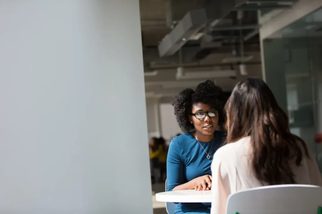 Duas mulheres, uma com óculos e cabelo encaracolado e a outra de costas, sentadas em uma mesa em um escritório, conversando. A imagem representa uma entrevista de desligamento ou uma conversa de trabalho.