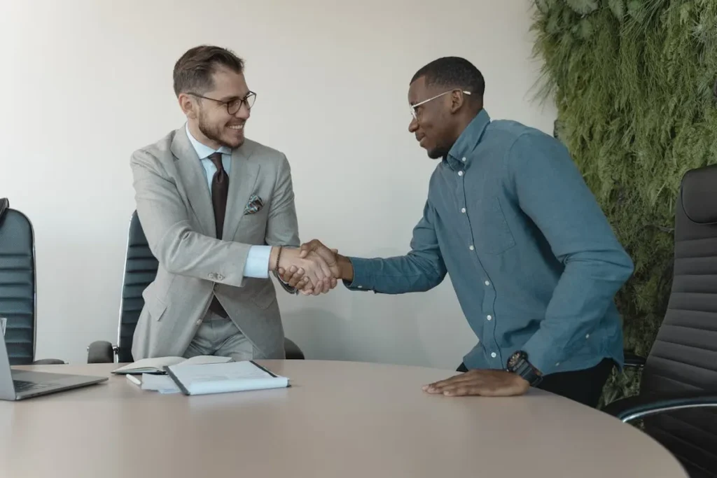 Dois homens, um com terno e outro com uma camisa casual, apertando as mãos em um escritório. A imagem destaca a importância da comunicação e da preparação para entrevistas em inglês.