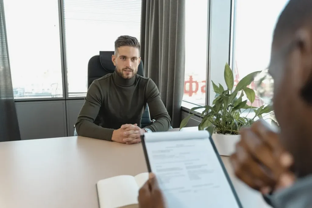 Um candidato em uma entrevista de emprego. Um homem de gola alta e barba está sentado em frente a uma pessoa que o entrevista, enquanto ele segura uma prancheta. A imagem destaca a importância da preparação para as entrevistas em inglês.