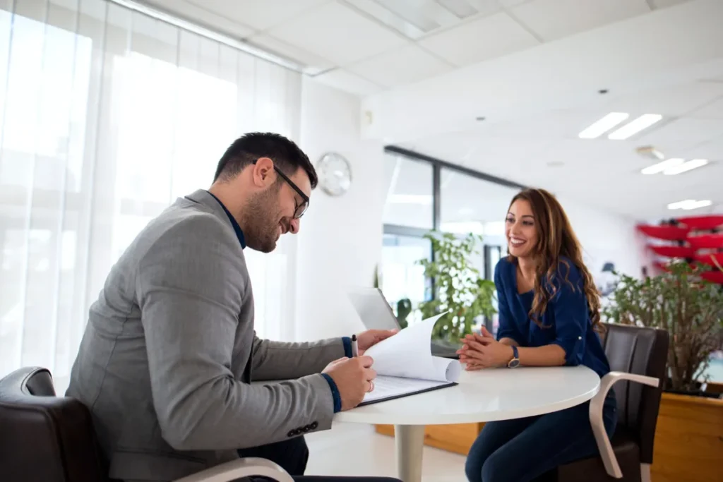 Um recrutador e uma candidata sorriem e conversam em uma entrevista de emprego, sentados em uma mesa redonda em um escritório moderno. O homem folheia um documento, enquanto a mulher escuta atentamente, em um ambiente amigável.