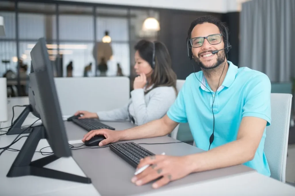 Um homem sorrindo com um headset e olhando para a câmera. Ele está sentado em uma mesa de escritório com um monitor e teclado. A imagem sugere a importância do inglês básico para o atendimento ao cliente e a comunicação profissional.