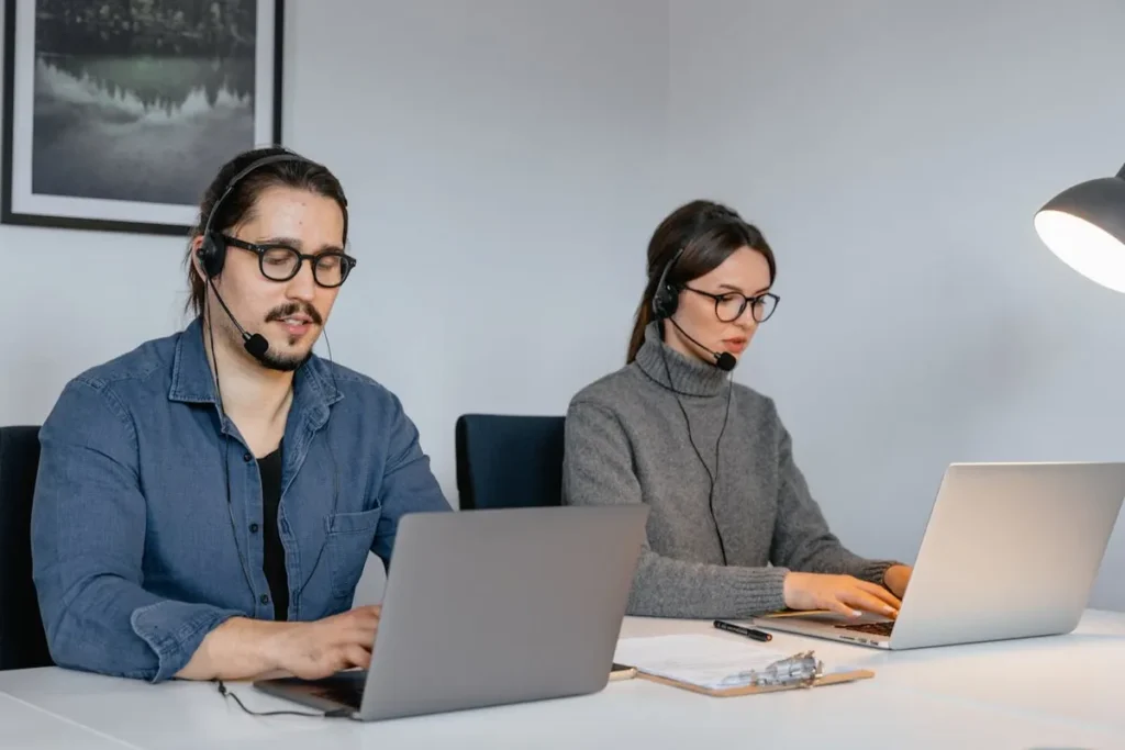 Dois atendentes de call center, um homem e uma mulher, usando headsets enquanto trabalham em seus laptops. A imagem destaca a necessidade de inglês básico para a comunicação profissional em empresas.