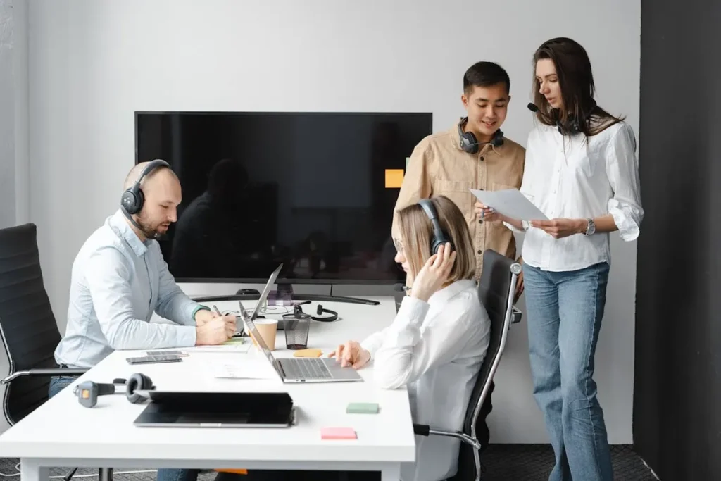 Pessoas trabalhando em um escritório moderno, usando fones de ouvido e laptops. A imagem destaca a importância da comunicação e do inglês básico para a colaboração em equipe.