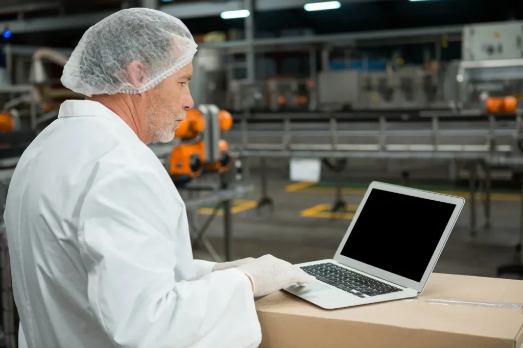 Engenheiro de produção com um gorro de cabelo e luvas, usando um laptop em uma caixa na linha de produção. A imagem foca na otimização, controle de qualidade e inspeção da produção.