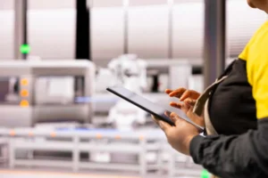 Pessoa com as mãos em um tablet, monitorando uma linha de produção automatizada ao fundo. A imagem destaca a tecnologia, controle de qualidade e a supervisão de operações industriais.