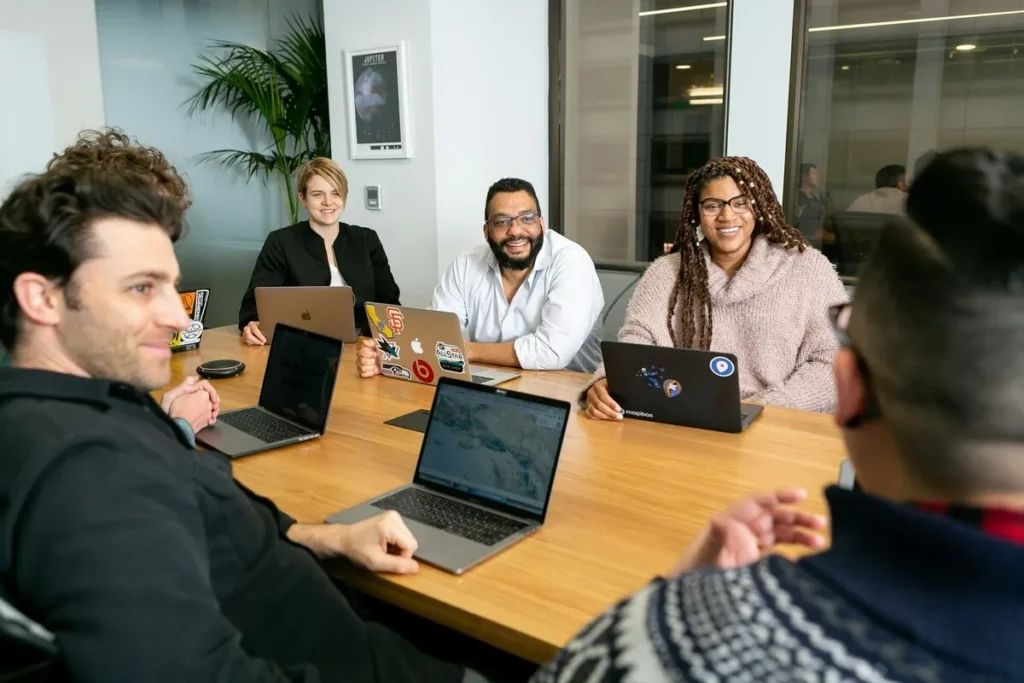 Uma equipe diversa de seis pessoas em uma reunião de onboarding no escritório, sentadas ao redor de uma mesa de madeira, com laptops abertos e sorrindo.