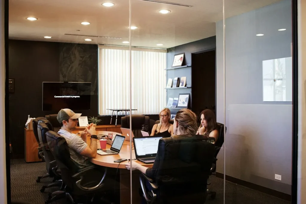 Quatro profissionais em uma reunião de onboarding em uma sala de conferências moderna, sentados ao redor de uma mesa oval com laptops abertos e discutindo estratégias.