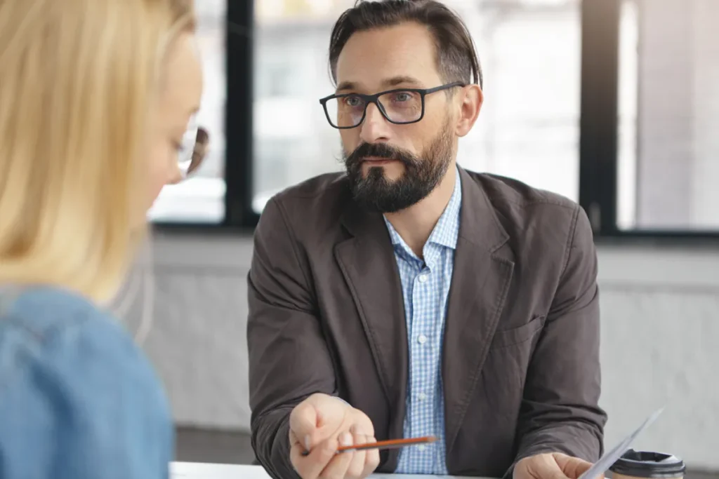 Um recrutador com barba e óculos, segurando uma caneta e um documento, faz perguntas em uma entrevista de emprego a uma candidata loira em um escritório. A imagem captura a interação profissional durante o processo seletivo.