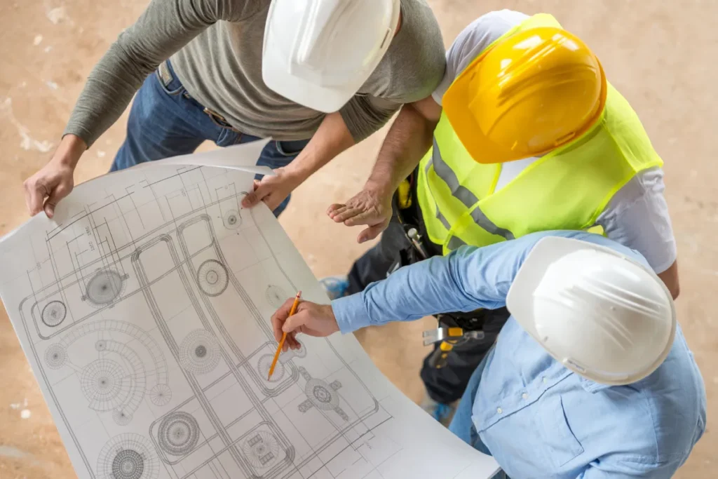 Dois profissionais com capacetes de segurança analisando uma planta técnica de engenharia sobre uma mesa.