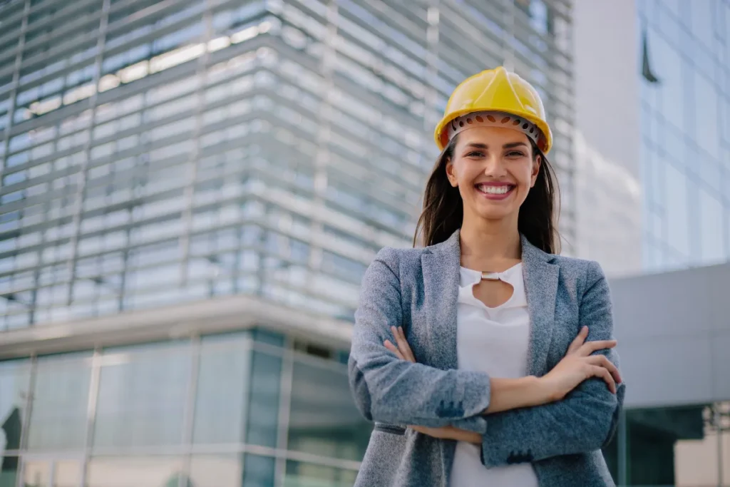 Engenheira usando capacete de segurança com os braços cruzados em frente a um prédio moderno.