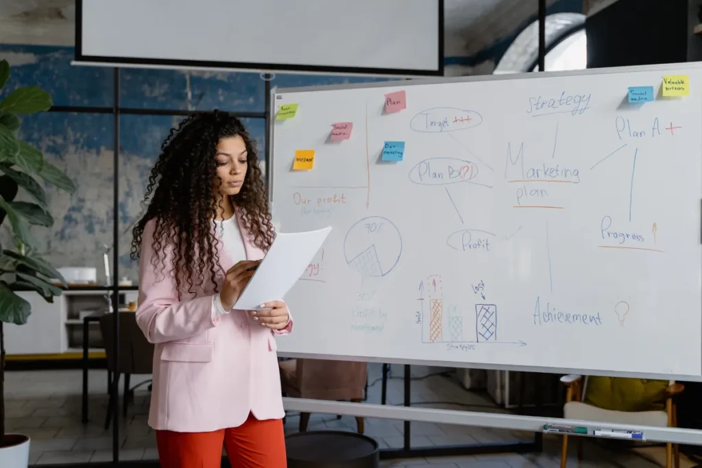 Mulher apontando para um quadro com anotações e diagramas durante uma apresentação em ambiente corporativo.