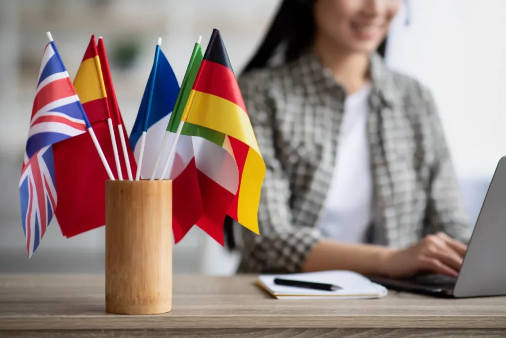 Pequenas bandeiras de diferentes países sobre uma mesa enquanto uma pessoa trabalha ao fundo em um ambiente de escritório.