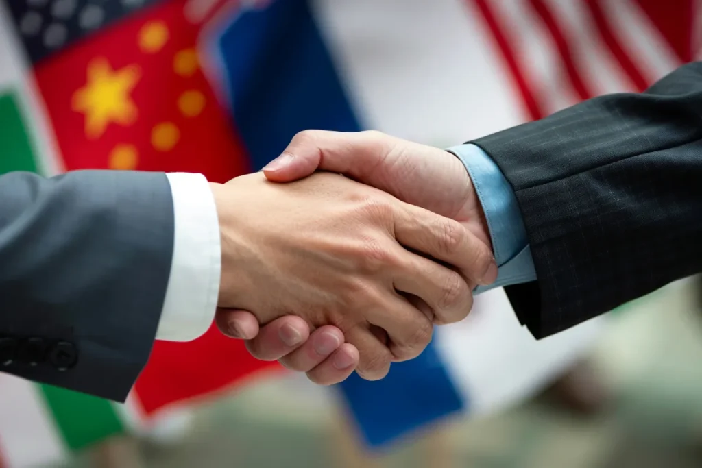 Duas pessoas apertando as mãos enquanto bandeiras de diferentes países aparecem desfocadas ao fundo.