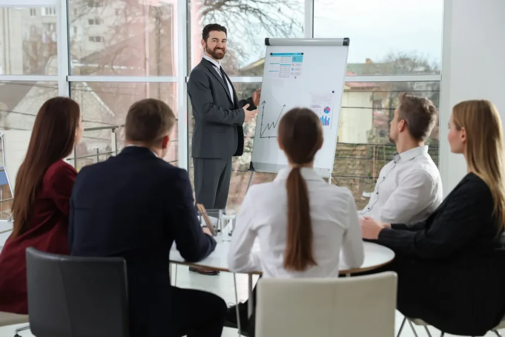 Profissional apresentando dados em uma tela enquanto um grupo de executivos acompanha a reunião em uma sala corporativa.