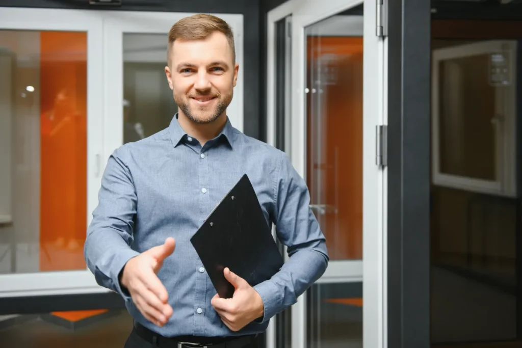Profissional de pós-venda sorrindo enquanto estende a mão para cumprimentar um cliente em ambiente corporativo, segurando uma prancheta.