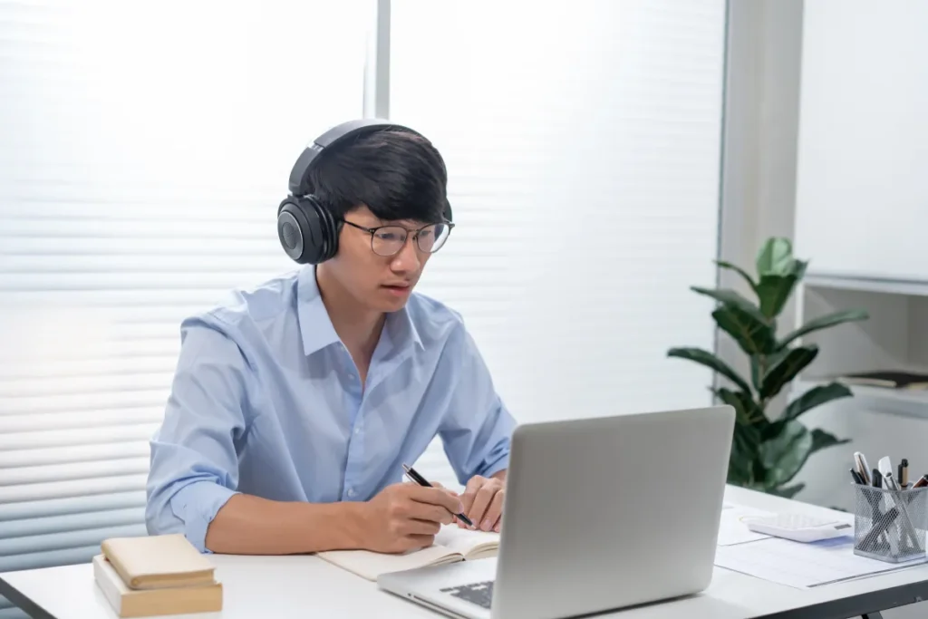 homem usando fones de ouvido estudando no notebook e fazendo anotações em um caderno na mesa de trabalho